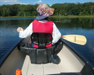 View from the back of a canoe on Clamshell Pond of the paddler in front and the surrounding pond and treeline