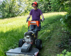 A volunteer wearing a safety hardhat and ear protectors walking behind a brush cutting machine mowing a grassland trail